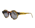 A pair of round full frame sunglasses with a patterned frame, likely to be displayed against a white background. Kuboraum Maske K9 tortoiseshell/amber-horn acetate round sunglasses; chunky sculptural beveled rims and wide temples with grey lenses; lightweight, comfortable, unisex; works with tailoring, streetwear, artwear, festivals; avant-garde/arthouse/Berlin design; brand born in Berlin, handmade in Italy, luxury indie eyewear, rare model.